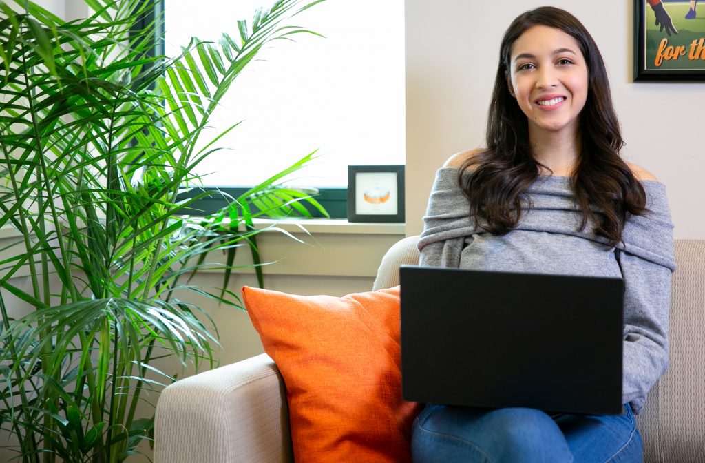 Female student sitting on couch with laptop smiling at camera.