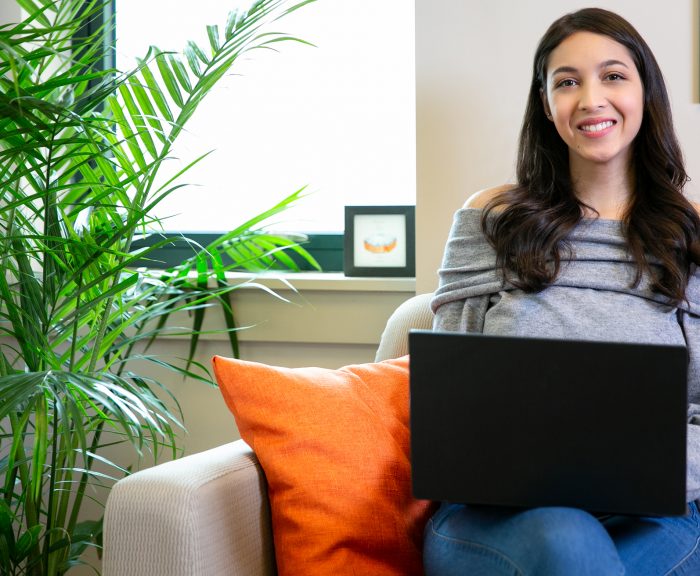 Female student sitting on couch with laptop smiling at camera.