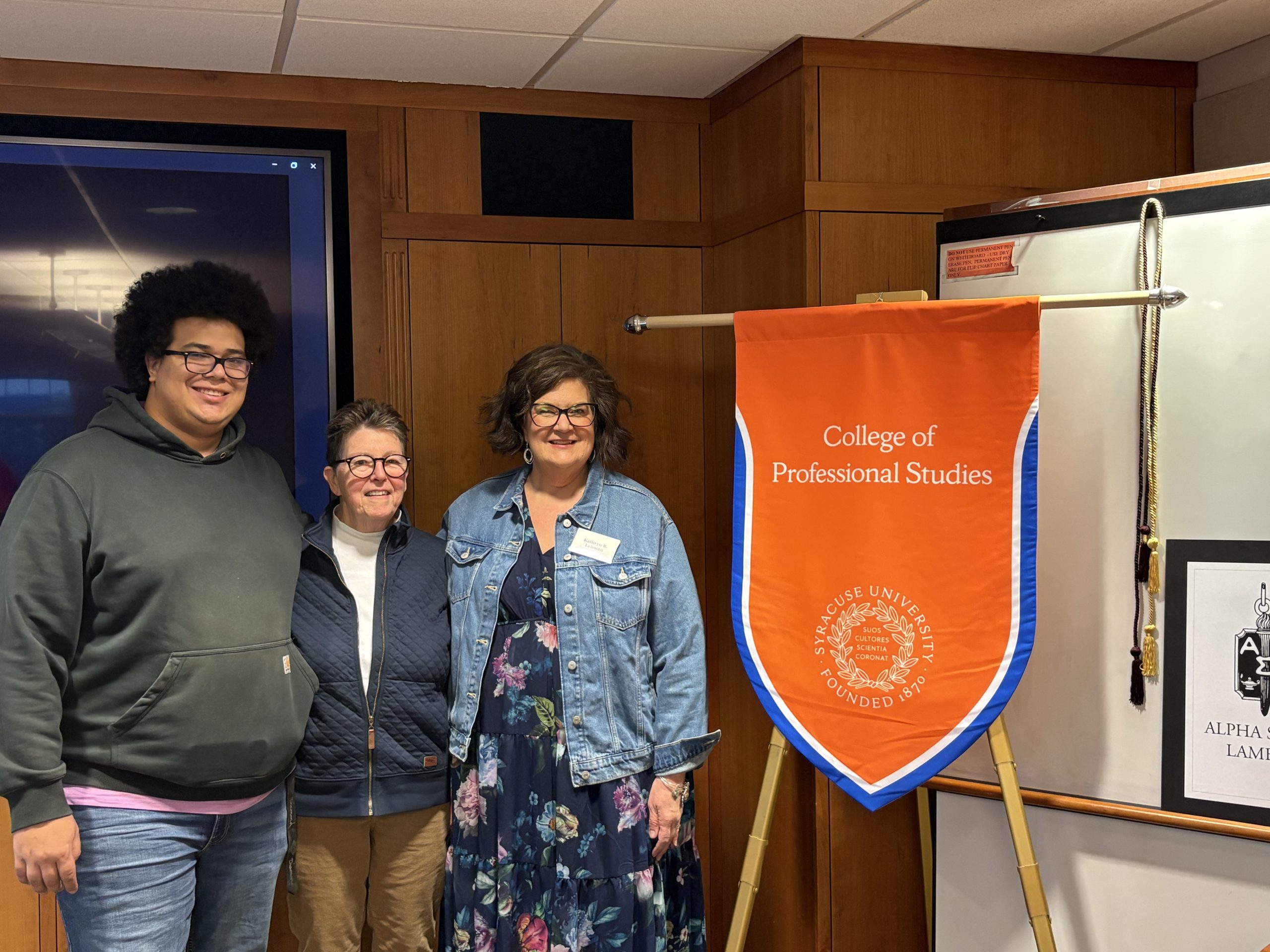 Three people stand next to the orange College of Professional Studies banner with white text. 