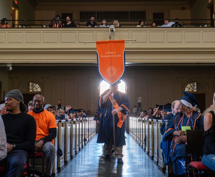 Graduate in blue gown carries an orange banner reading 'College of Professional Studies' down the aisle at a graduation ceremony.
