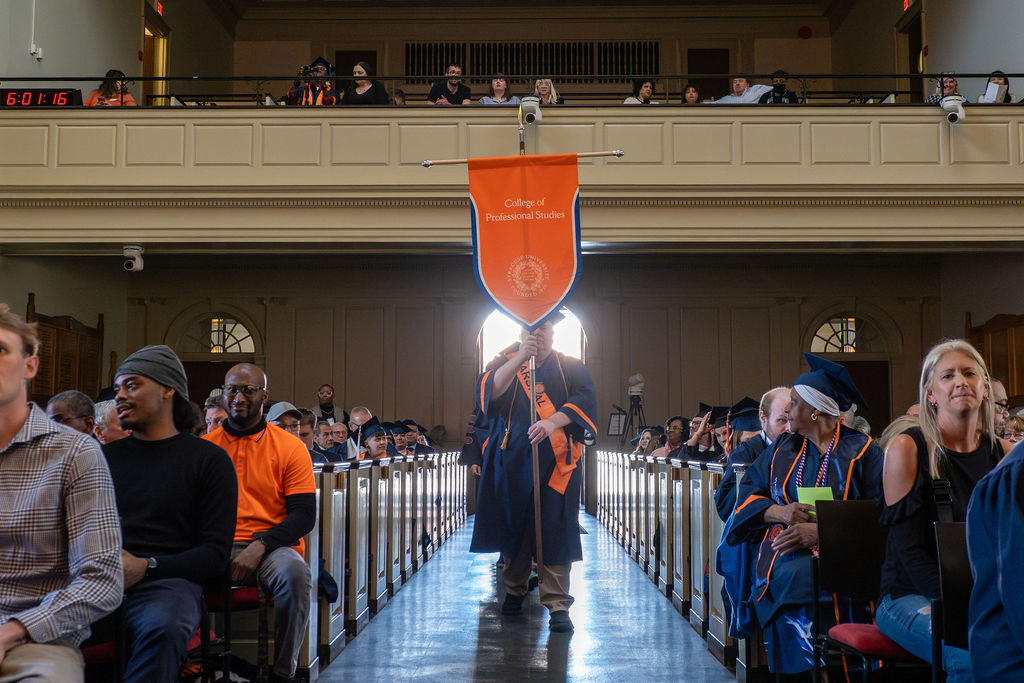 College Marshal, Kristopher Plantz, carries a banner that says "College of Professional Studies" down an aisle. 