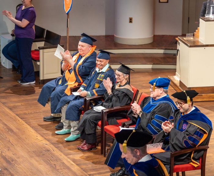 Faculty and graduates in caps and gowns seated on stage, clapping during a graduation ceremony.